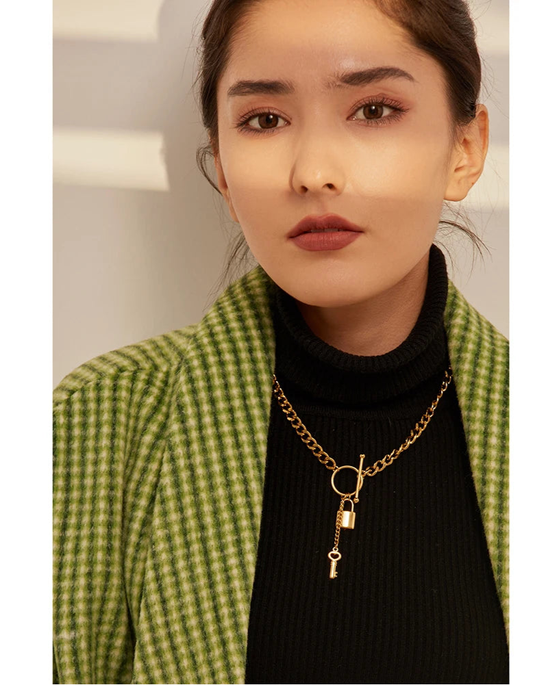 Woman wearing a green checkered coat and black turtleneck with a gold necklace featuring a key pendant.