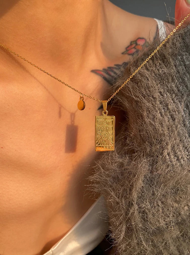 Gold zodiac necklace with a rectangular zodiac pendant worn by a person with tattoos on their neck.