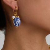 Gold earring with a blue and white floral design on a person's ear against a neutral background