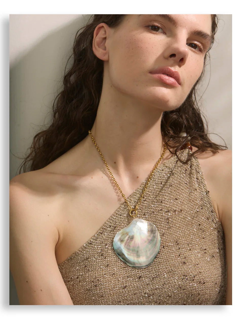 Woman wearing a gold necklace with a large shell pendant against a neutral background