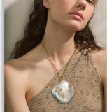 Woman wearing a gold necklace with a large shell pendant against a neutral background