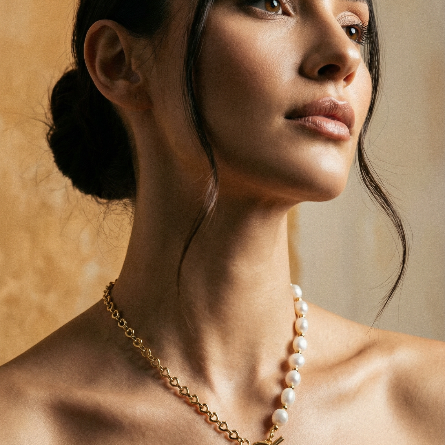 Woman wearing a gold necklace with a heart pendant against a warm-toned background