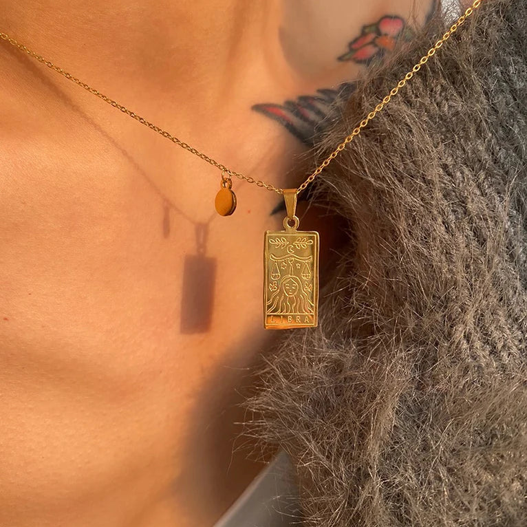 Gold zodiac necklace with a rectangular zodiac pendant worn by a person with tattoos on their neck.