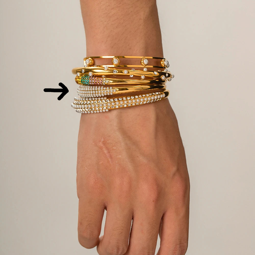 Hand wearing multiple gold and pearl bracelets on a neutral background

