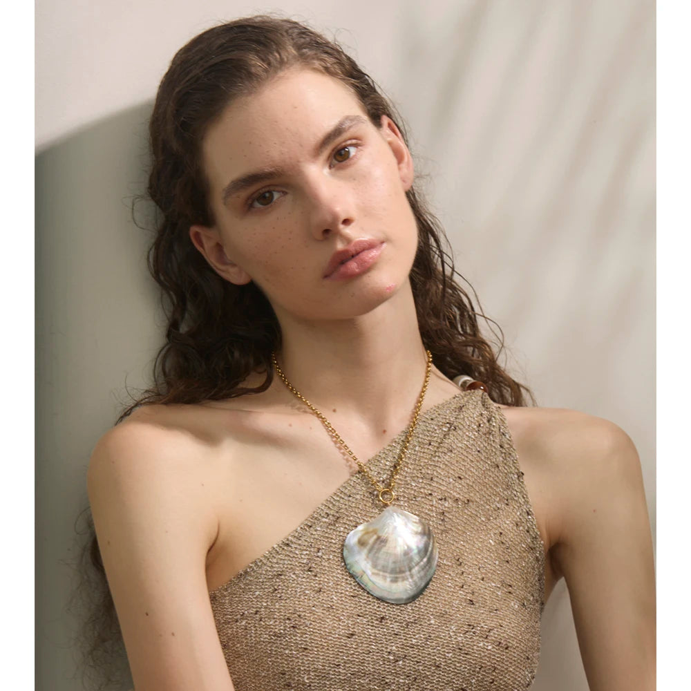 Woman wearing a gold necklace with a large shell pendant against a neutral background