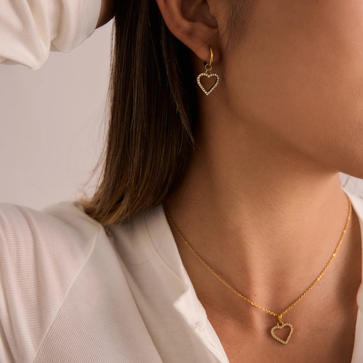 Close-up of a person wearing gold heart-shaped earrings and necklace against a neutral background