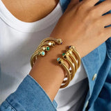 A close-up of a person's wrist wearing multiple gold-toned rope bracelets with small colorful gemstone accents, styled with a white ribbed top and a blue denim jacket.