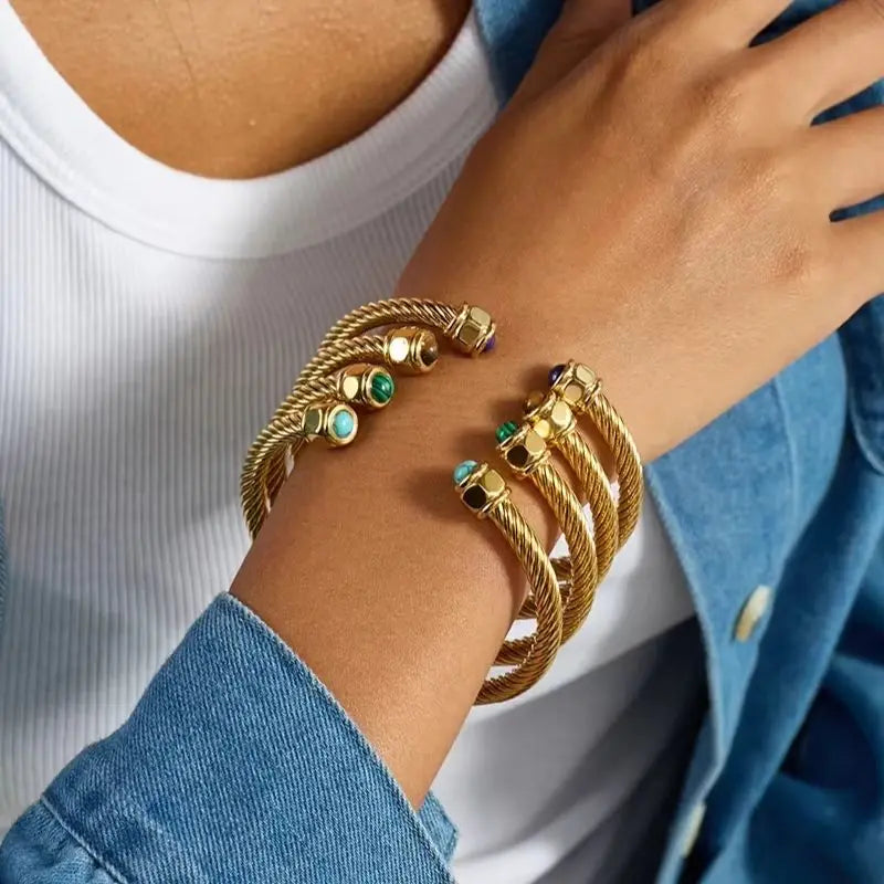 A close-up of a person's wrist wearing multiple gold-toned rope bracelets with small colorful gemstone accents, styled with a white ribbed top and a blue denim jacket.