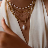Close-up of layered gold necklaces with pearl pendants on a person wearing a white top.