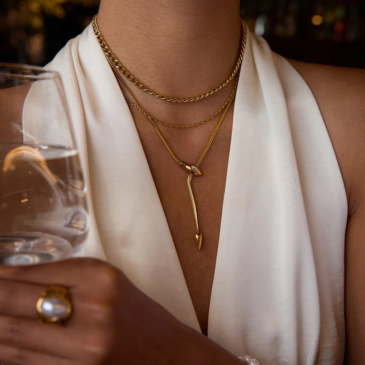 Person wearing gold necklaces and a ring, holding a glass with a blurred background
