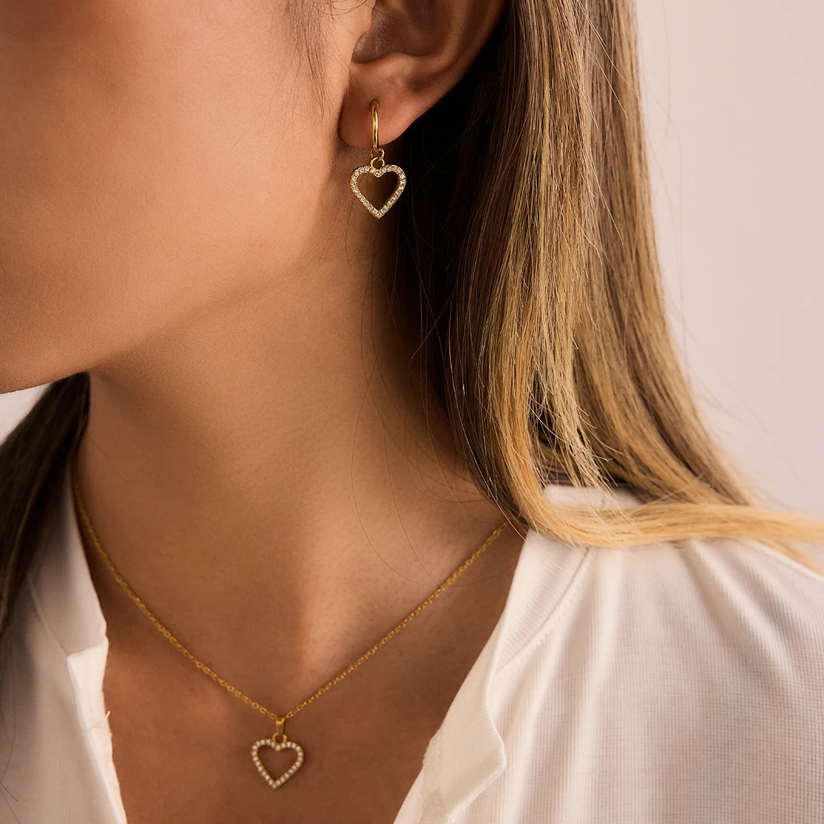 Close-up of a person wearing gold heart-shaped earrings and necklace against a neutral background